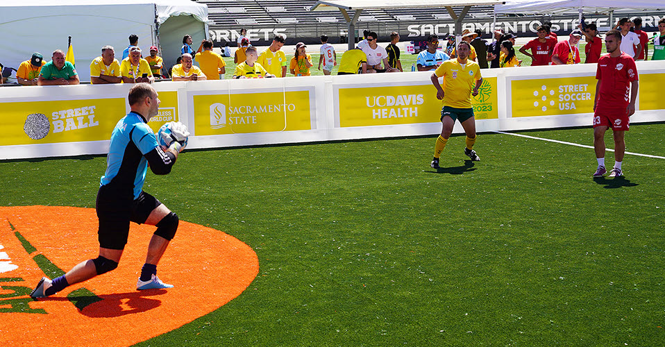 Athletes play on a football (soccer) pitch during the 2023 Homeless World Cup in Sacramento, California. The athlete on the left is standing in front of the goal and holding the ball, wearing a blue and black uniform. Two athletes on the right are watching, one in a yellow jersey and the other in red.