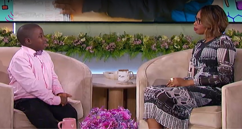 Sean Atitsogbe, a little Black boy wearing a pink button-down shirt and black pants is sitting in a chair opposite Jennifer Hudson, who is wearing a long printed and pattern dress, discussing Sean's love of science on an episode of The Jennifer Hudson Show.